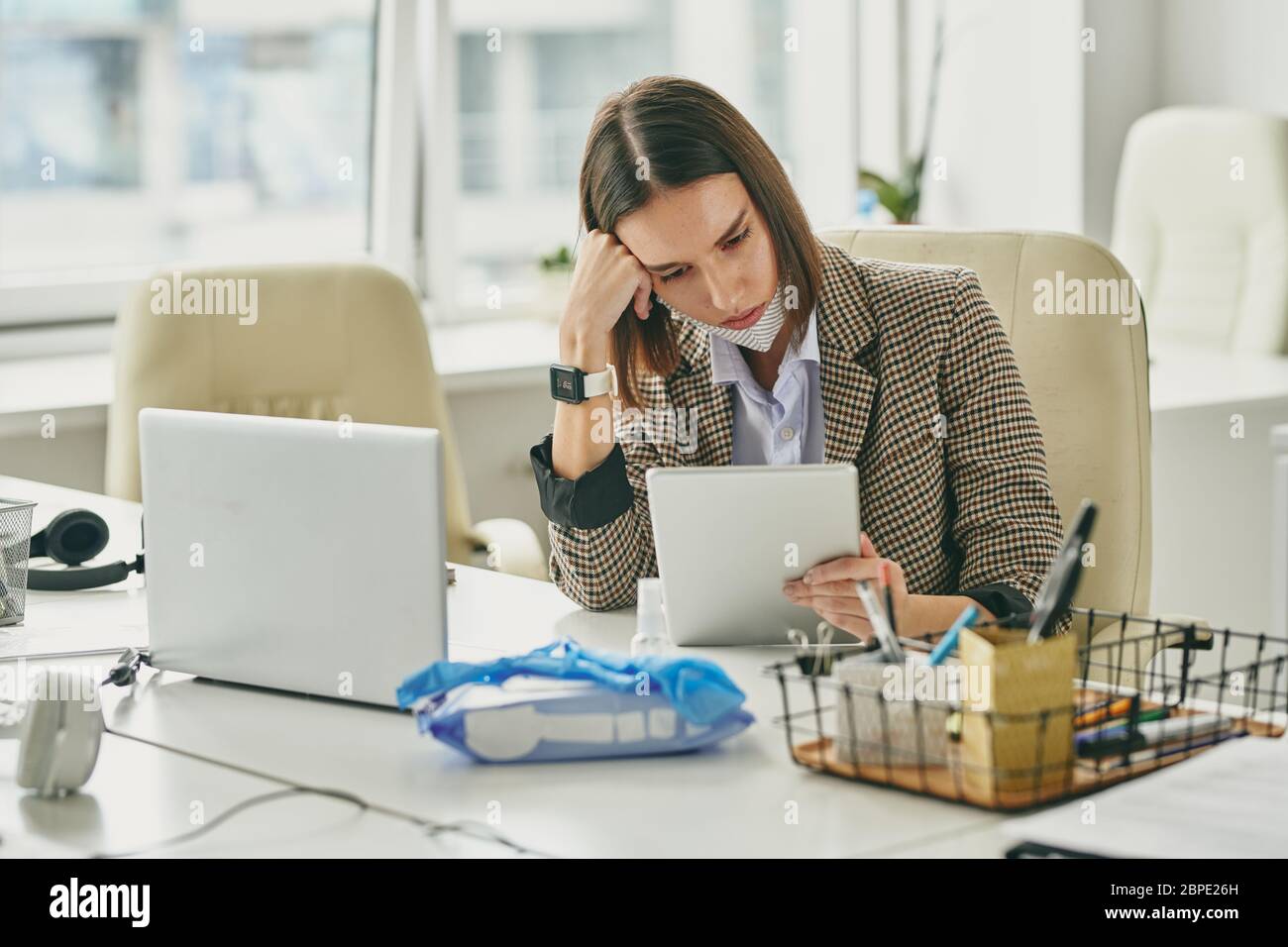Seriöse müde junge Geschäftsfrau mit Maske auf Kinn sitzt am Schreibtisch mit Laptop und überprüfen Sie die Post auf Tablet im leeren Büro während der covid-19 Stockfoto