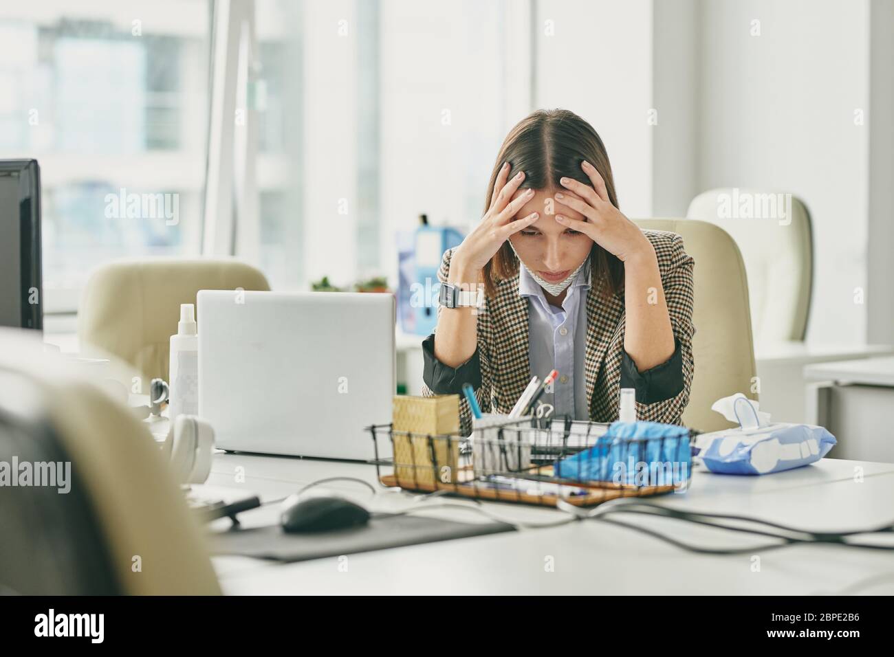 Verwirrt junge Bürofrau sitzt am Schreibtisch in leerem Büro während Coronavirus-Epidemie und hält Kopf in den Händen, während Probleme Stockfoto