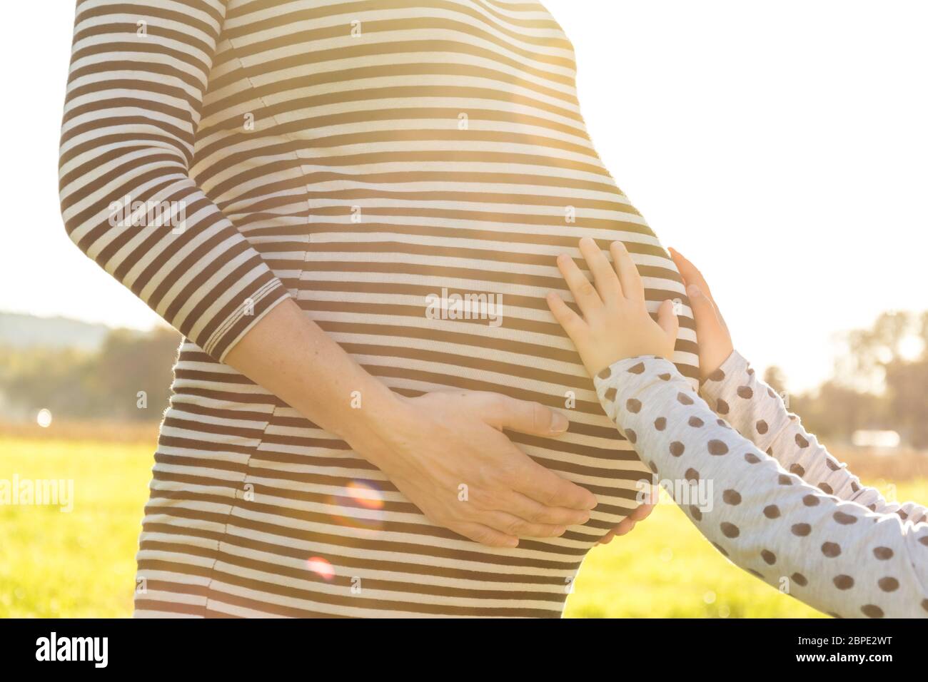Kleines Kind, das den schwangeren Bauch ihrer Mutter in malerischer Nachmittagssonne berührt Stockfoto