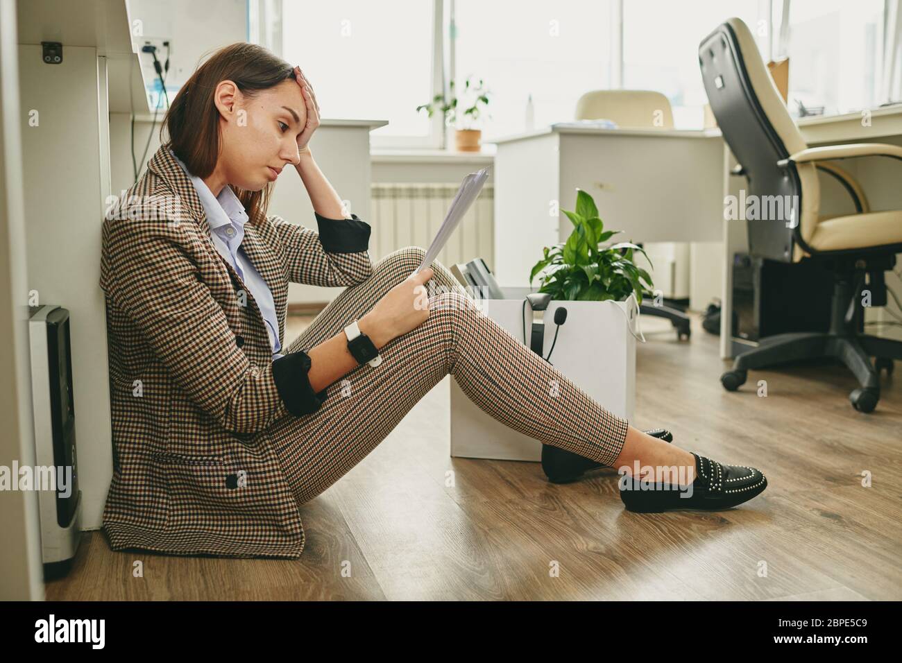Verärgert junge Frau in stilvollen Casual Anzug sitzen auf dem Boden und lesen Hinweis auf Entlassung in leerem Büro während Coronavirus Quarantäne Stockfoto