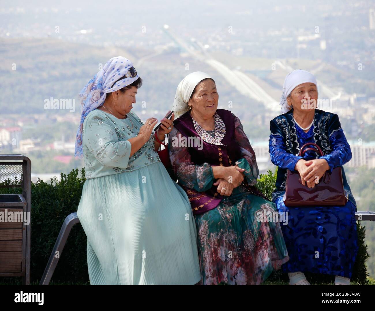 Almaty, Kasachstan - 3. September 2019: Drei ältere Frauen ruhen sich auf einem Aussichtspunkt in Almaty aus, während eine von ihnen ihr Mobiltelefon benutzt. Stockfoto