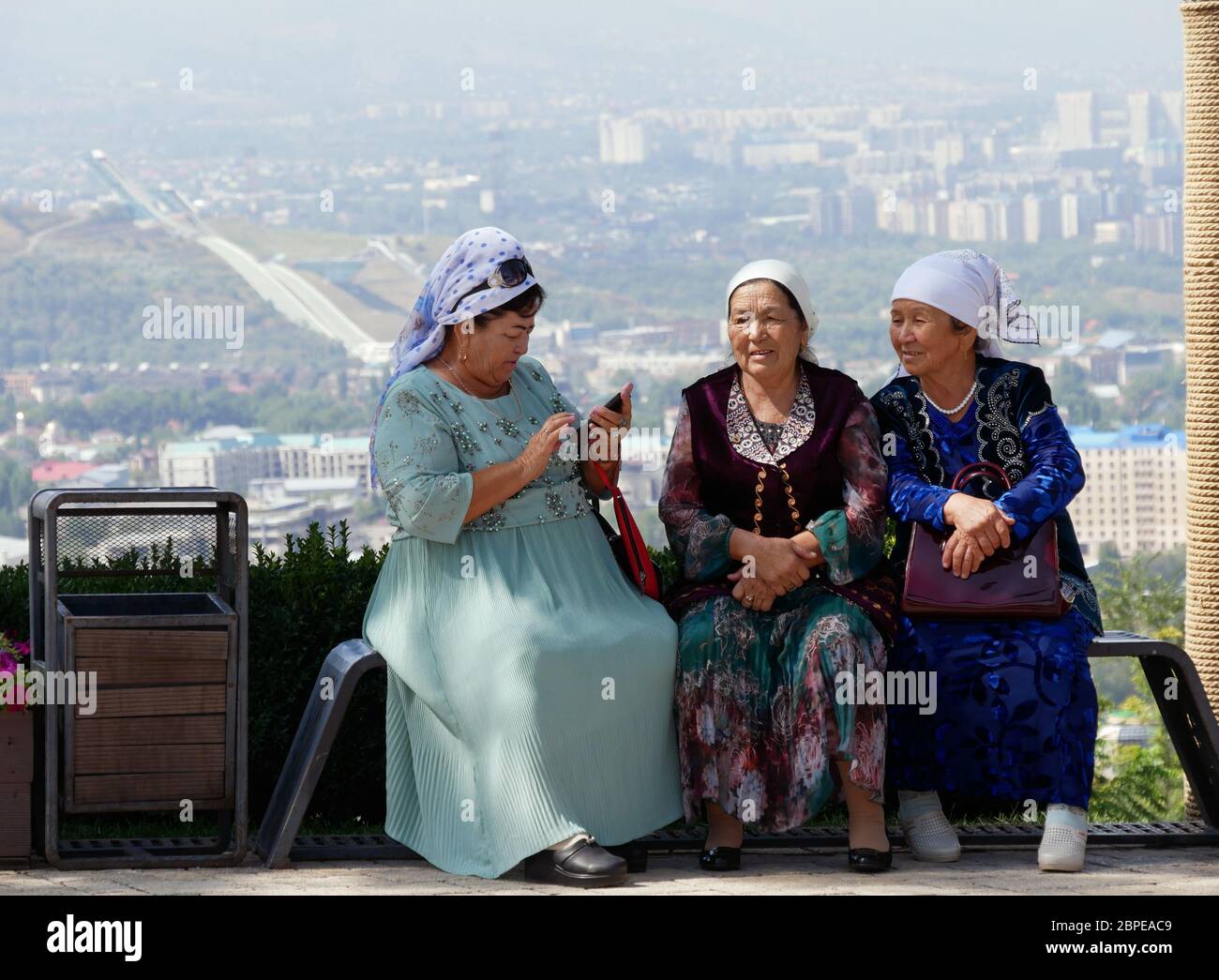 Almaty, Kasachstan - 3. September 2019: Drei ältere Frauen ruhen sich auf einem Aussichtspunkt in Almaty aus, während eine von ihnen ihr Mobiltelefon benutzt. Stockfoto