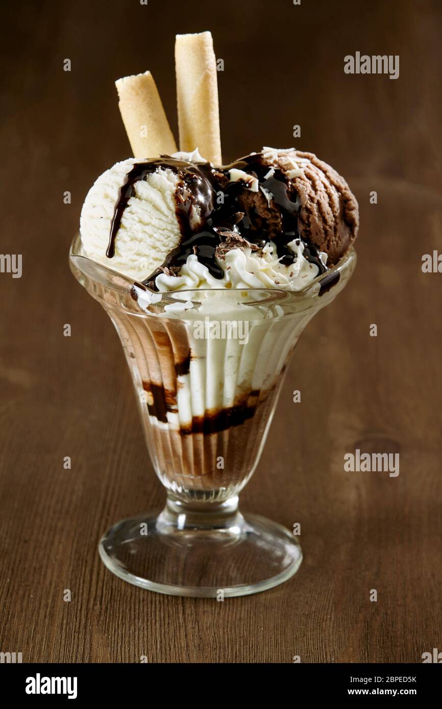 Köstliche Vanille- und Schokoladeneiskerze mit Schokoladensauce und Waffelkeksen in einem hohen Glas auf einer Holztheke serviert, Nahaufnahme Seite vie Stockfoto