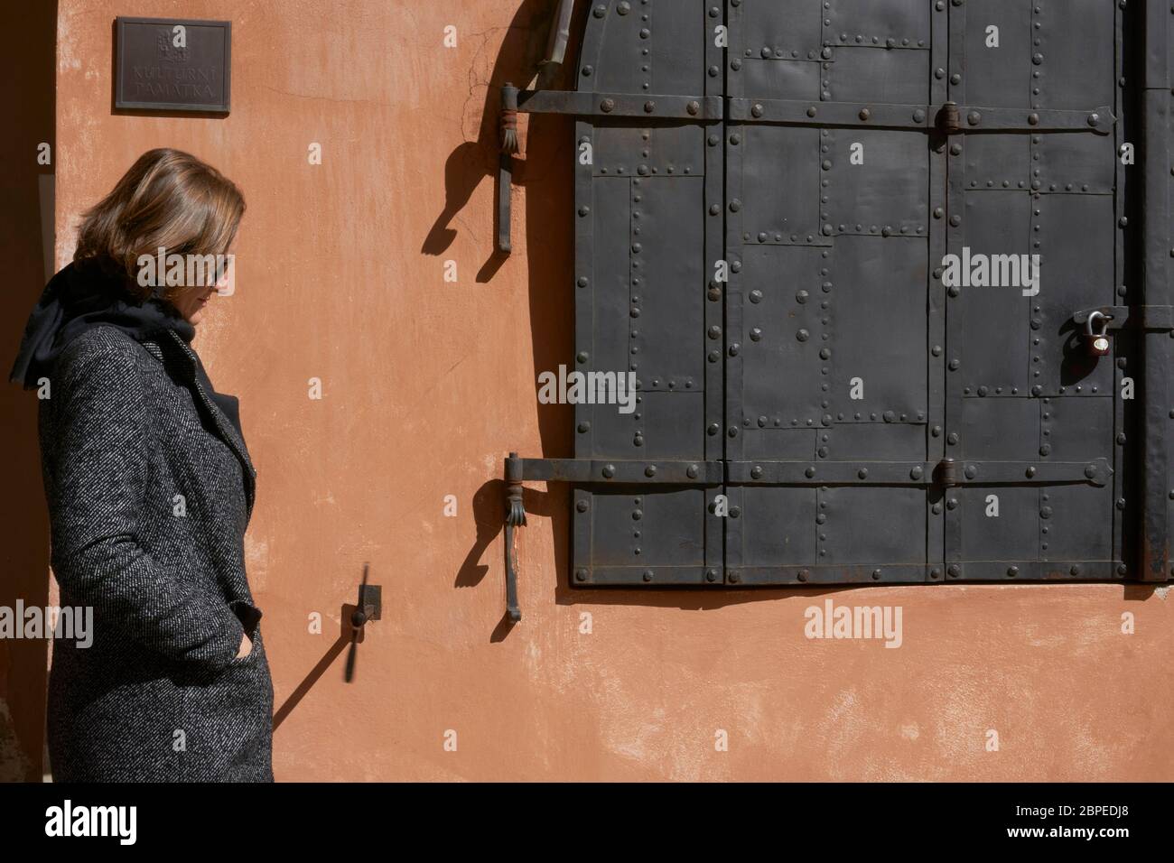 Eine Frau in einem grauen Mantel, die seitlich zur Kamera steht vor einem mittelalterlichen Haus mit orangefarbener Fassade und schwerem grauen Metallfenster Stockfoto