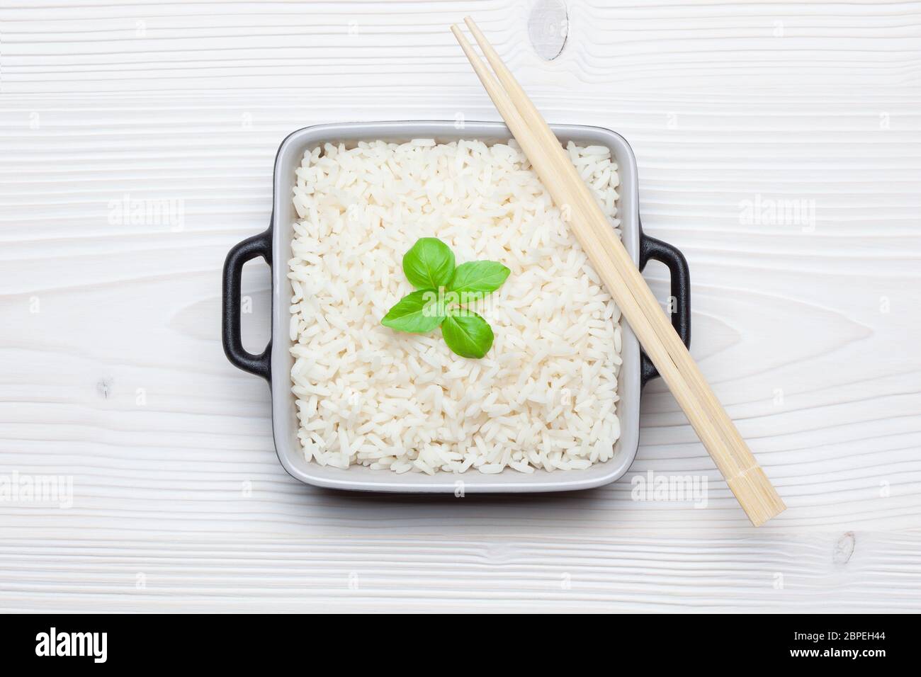 Reis mit Basilikum in Keramikschale mit Essstäbchen auf weißem Holztisch. Draufsicht. Stockfoto