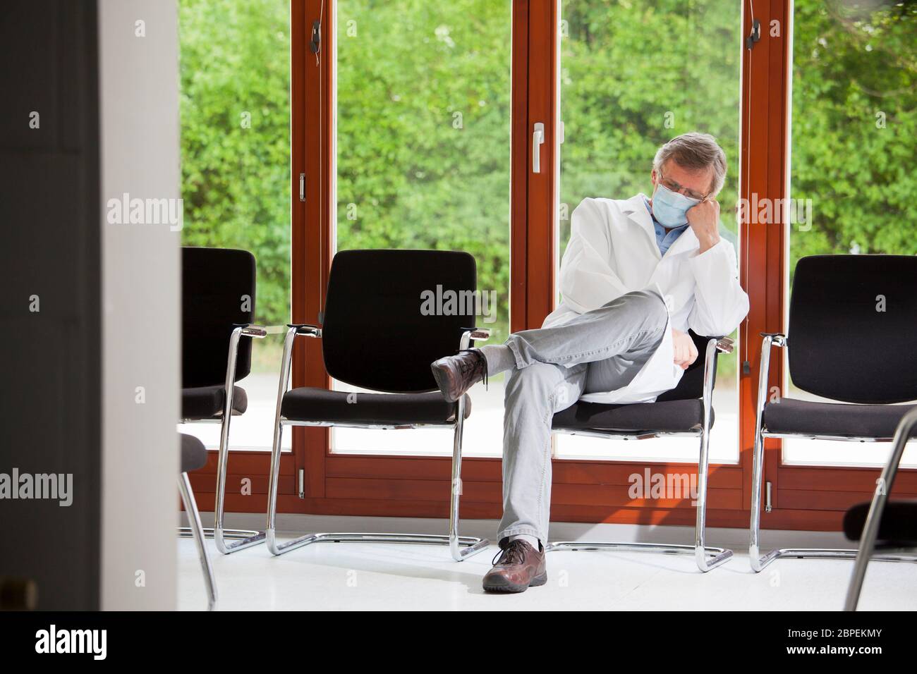 Überarbeiteter Arzt mit Gesichtsmaske, der in einem Stuhl in einem leeren Wartezimmer schläft - grüner Hintergrund mit Pflanzen Stockfoto