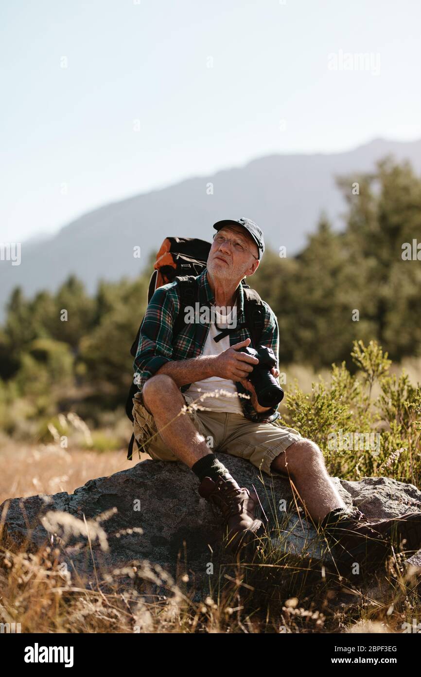 Älterer Mann auf Wandertour sitzt auf einem Felsen mit einer Digitalkamera. Reifer Wanderer, der mit einer Kamera in der Hand wegschaut. Stockfoto