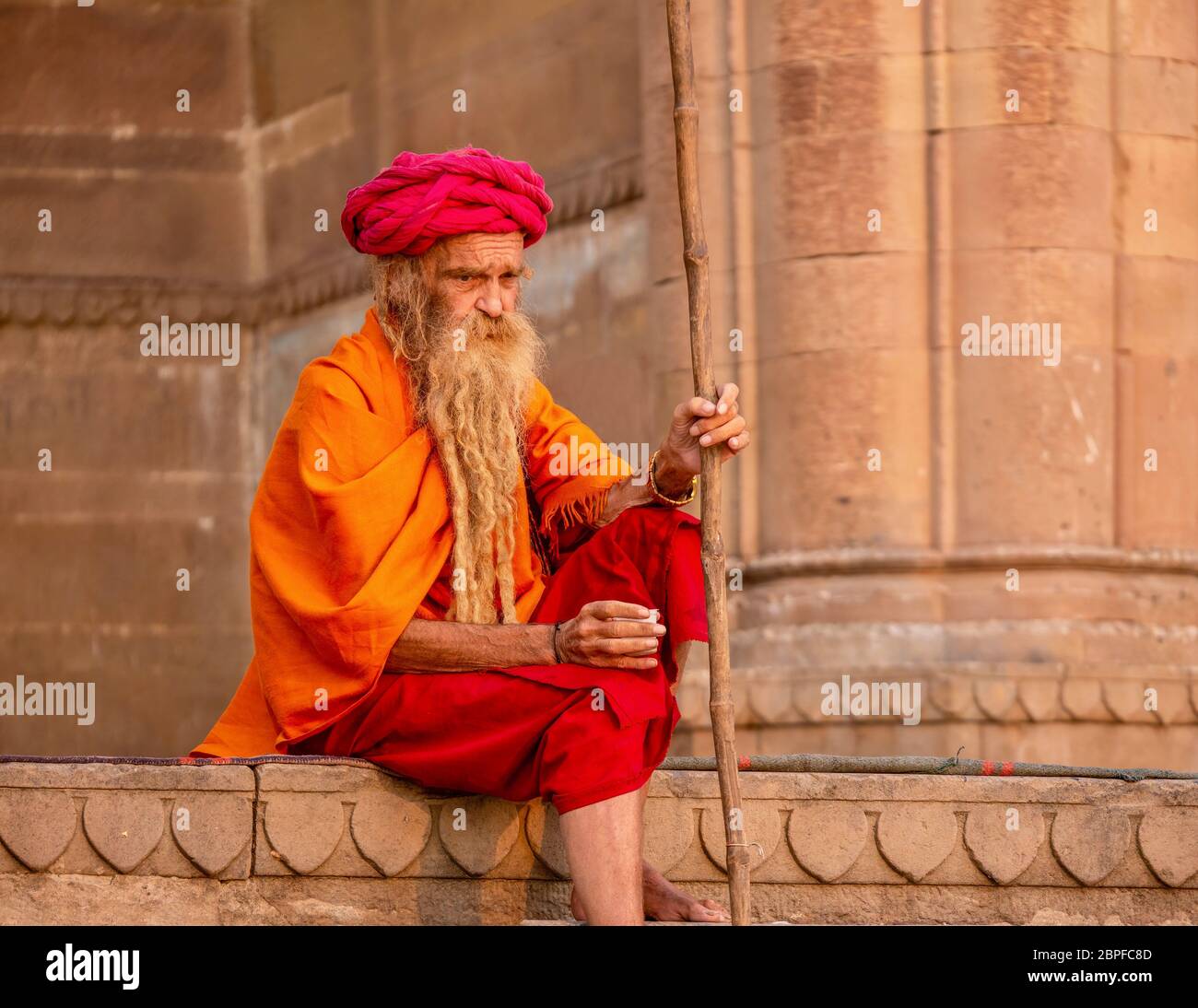 Varanasi, Indien - 13. November 2015. Ein kaukasischer Mann, der die bunte traditionelle Kleidung und den langen Bart eines Hindu-Sadhu trägt, sitzt auf den Ghats. Stockfoto