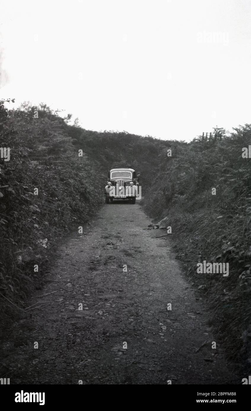 1948, historisch, geparkt auf einer schmalen, holprigen Landstraße oder Zufahrtsstraße zu Ackerland, umgeben von dicken, hohen Hecken, einem Ford Anglia, ein beliebtes Auto in dieser Zeit. In der Ferne kann man eine Dame sehen, die hinter dem Auto steht oder sich vielleicht versteckt, England, Großbritannien. Stockfoto