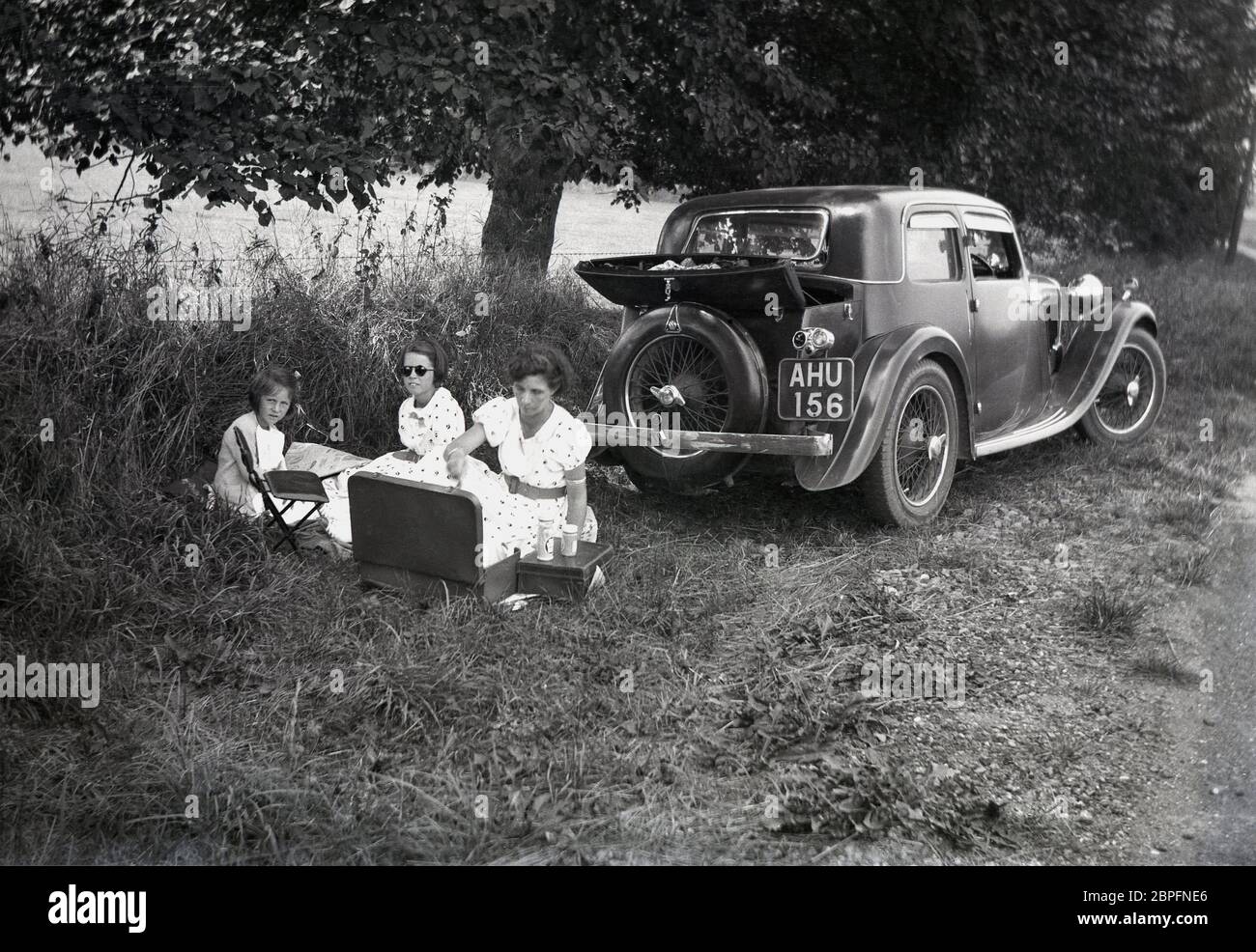 Auf einer kleinen Wiese, an einer Landstraße, sitzt eine Mutter am Heck eines Motorwagens mit Drahträdern, einem Sportlimousine der damaligen Zeit, und macht ein Picknick mit ihren beiden jungen Mädchen in Sommerkleider, circa 1940er Jahre. Stockfoto