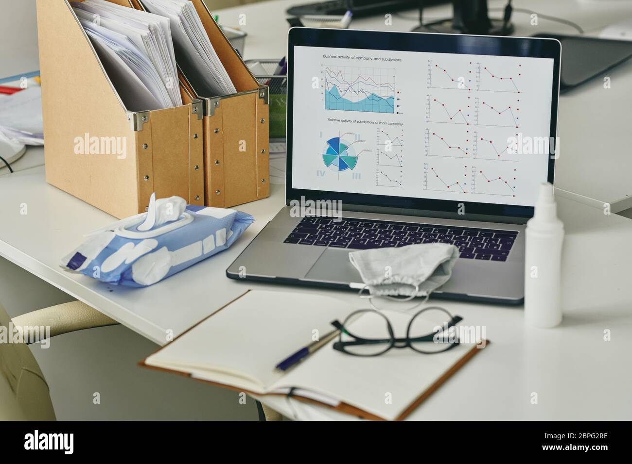 Abnehmende Diagramme auf Laptop-Bildschirm, Maske, Flasche antiseptisch, Paket von feuchten Wischtüchern auf dem Tisch des Büroangestellten Stockfoto