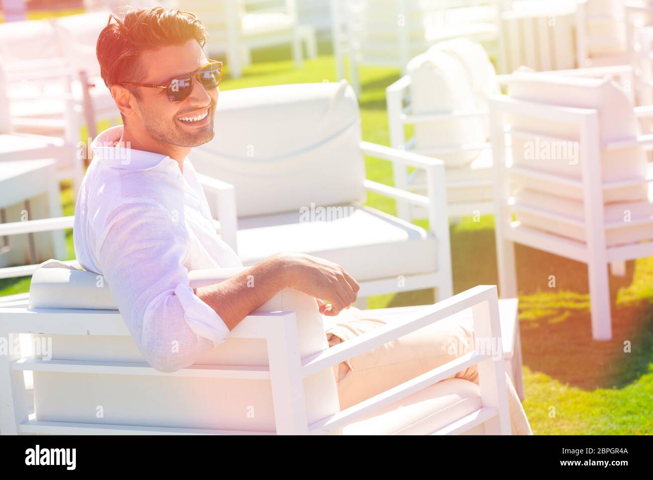 Fröhlicher und lächelnder Mann im Urlaub, der in einem Strandresort mit grüner Wiese sitzt. Mit Sonnenbrille. Intensives Tageslicht, trägt der junge Mann ein weißes Hemd und Stockfoto