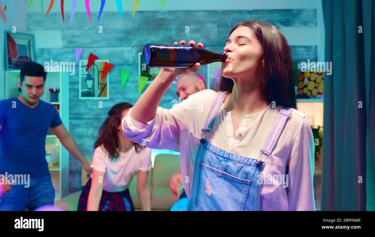 Zoom in Schuss von schönen Mädchen Bier aus der Flasche trinken auf wilden Party mit Neonlicht. Gruppe von Menschen tanzen im Hintergrund Stockfoto