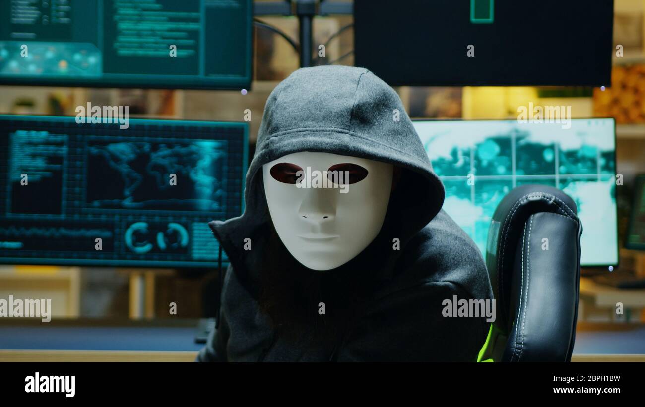 Maskierter Hacker in seiner Wohnung, der in die Kamera schaut und dabei Online-Informationen stiehlt. Stockfoto