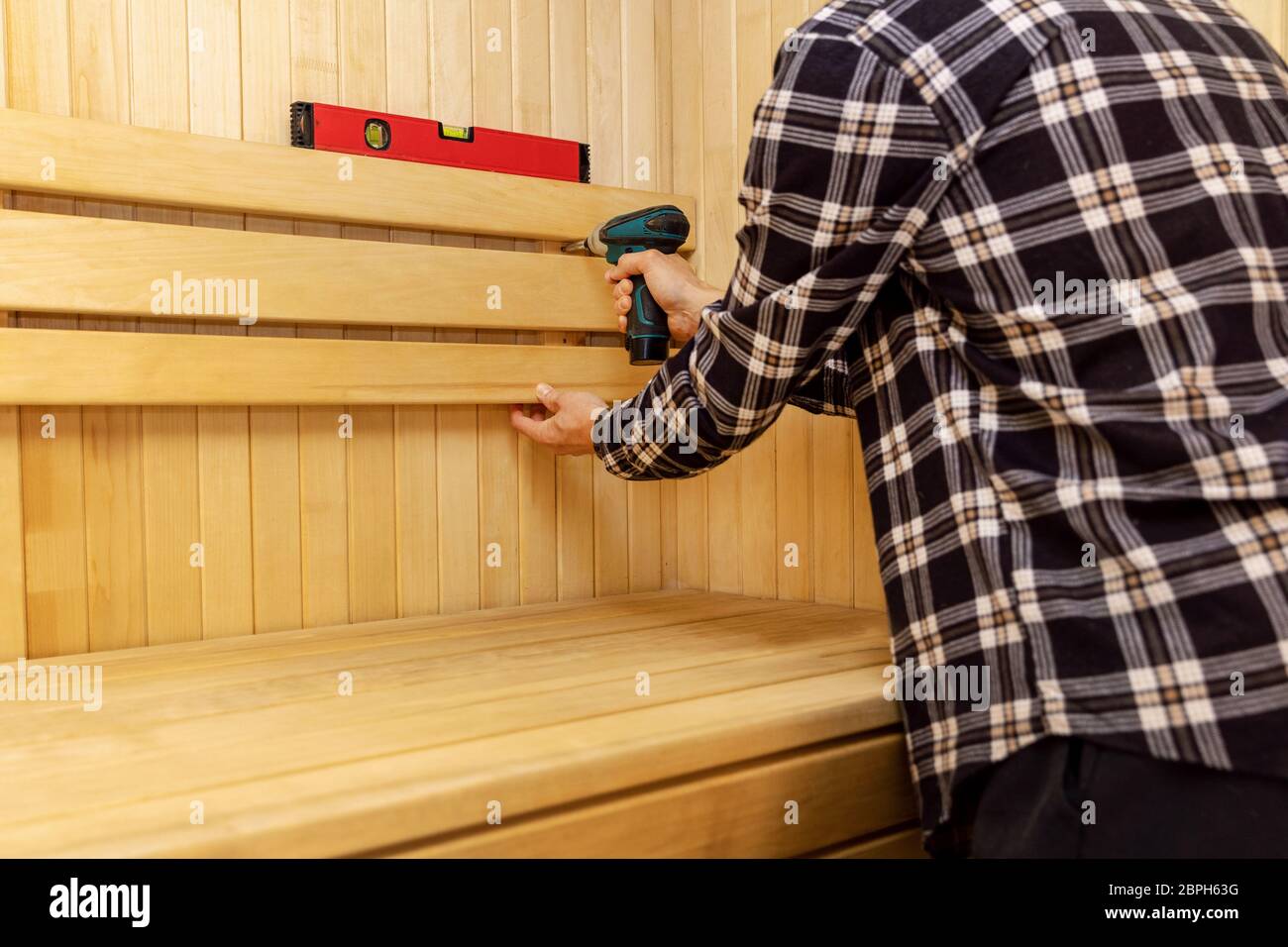 Sauna Bau - Mann verschraubt Holzbank Rückenlehne an der Wand Stockfoto