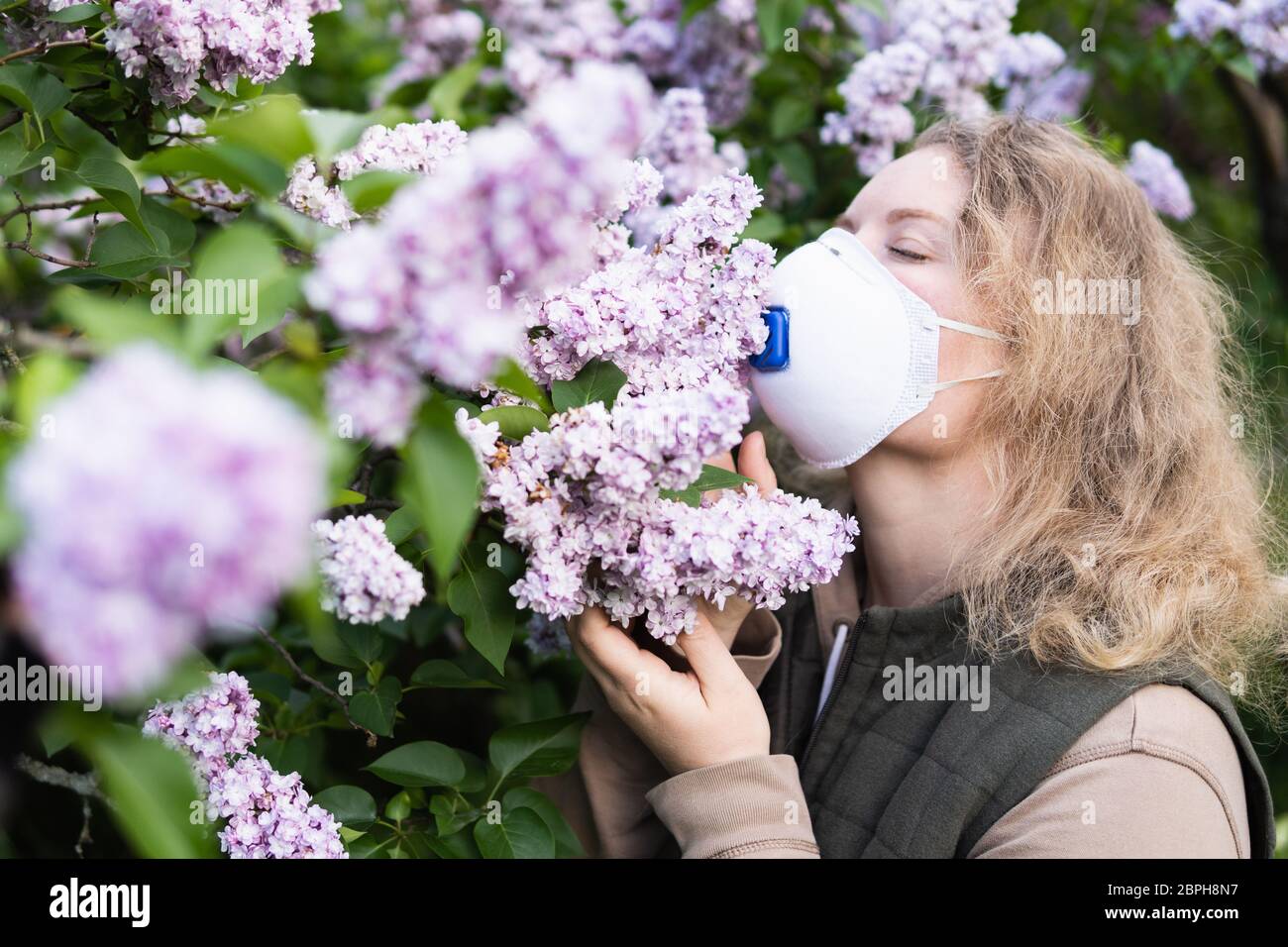 Junge blonde Frau riecht Flieder Baum Blüte durch FFP2 Atemschutzmaske. Konzeptionelles Bild der Quarantäne und Allergie des Corona-Virus. Stockfoto