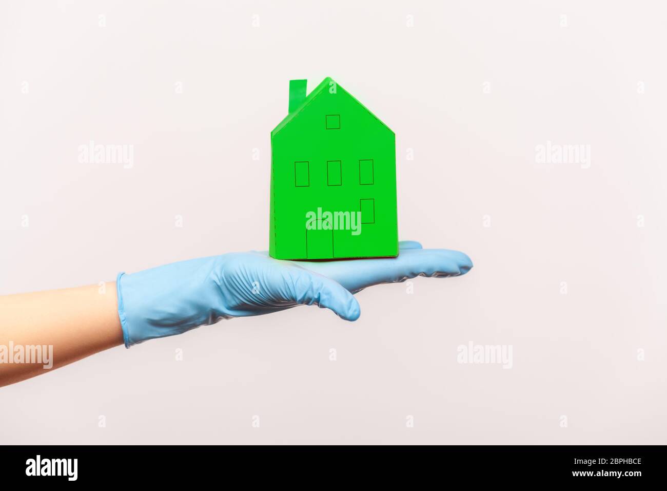 Profil Seitenansicht Nahaufnahme der menschlichen Hand in blauen chirurgischen Handschuhen, die grüne Papier Haus außen in der Hand. Indoor, Studio-Aufnahme, isoliert auf grauen Rücken Stockfoto