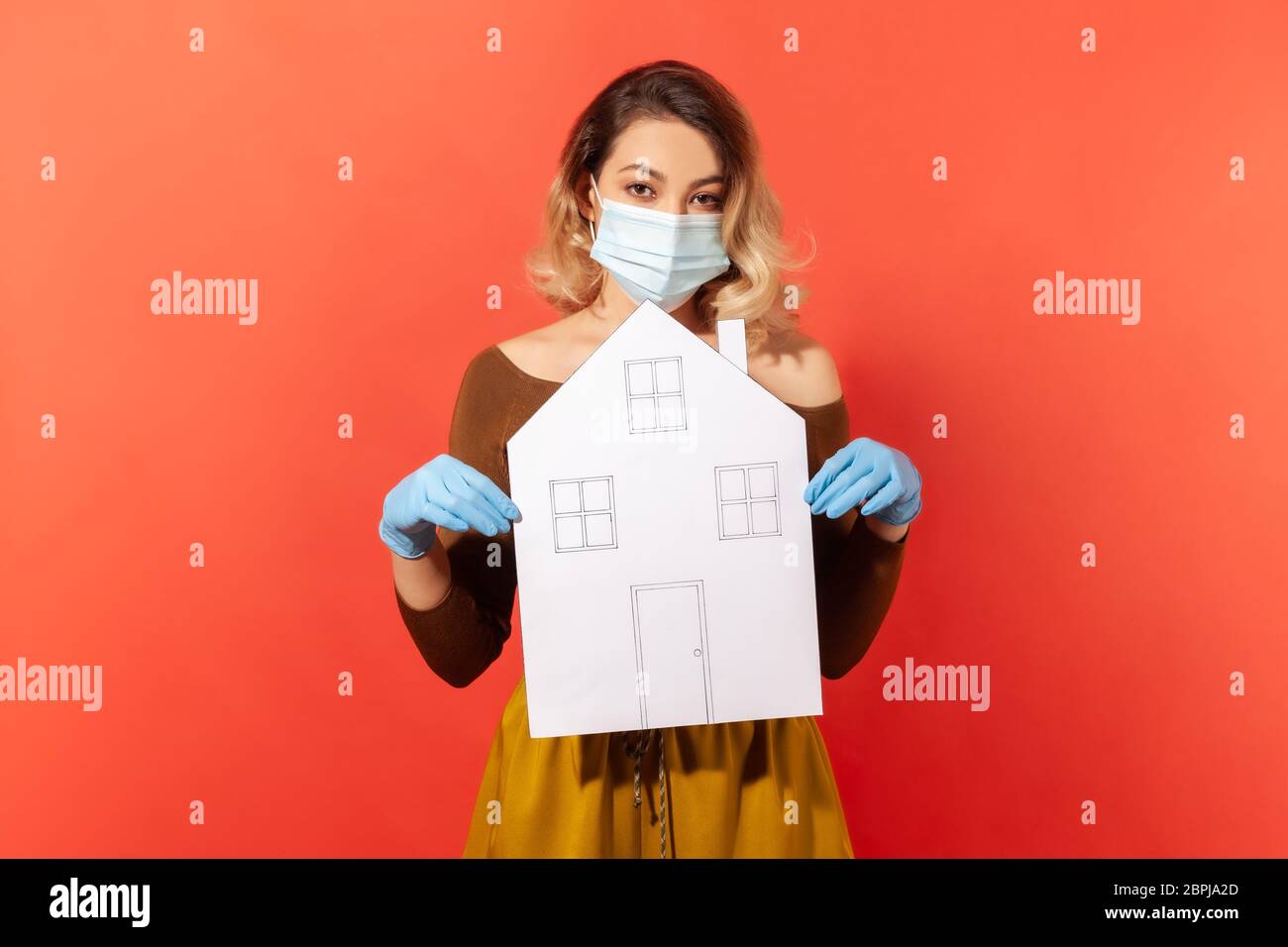 Junge Frau in chirurgischen Gesichtsmaske und Handschuhe halten Papierhaus, Empfehlung zu Hause Quarantäne bleiben, während ansteckende Krankheit Ausbreitung, coronavir Stockfoto
