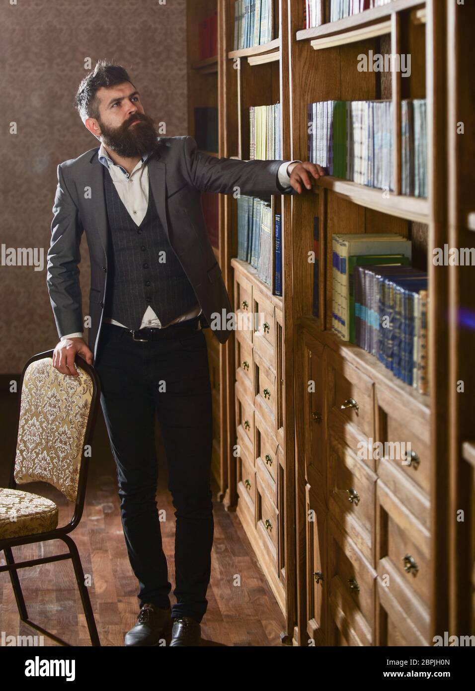 Intellektuelles Elite-Konzept. Aristokrat auf der Suche nach einem Buch. Mann in klassischen Anzug stehen in Vintage-Interieur, Bibliothek, Bücherregale auf Hintergrund. Altmodischer Mann, der Bücher anschaut, sucht. Stockfoto