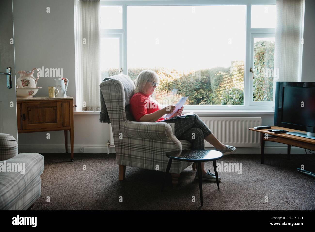 Eine ältere Frau sitzt in einem Sessel im Wohnzimmer ihres Hauses, Lesen durch Schreiben. Stockfoto