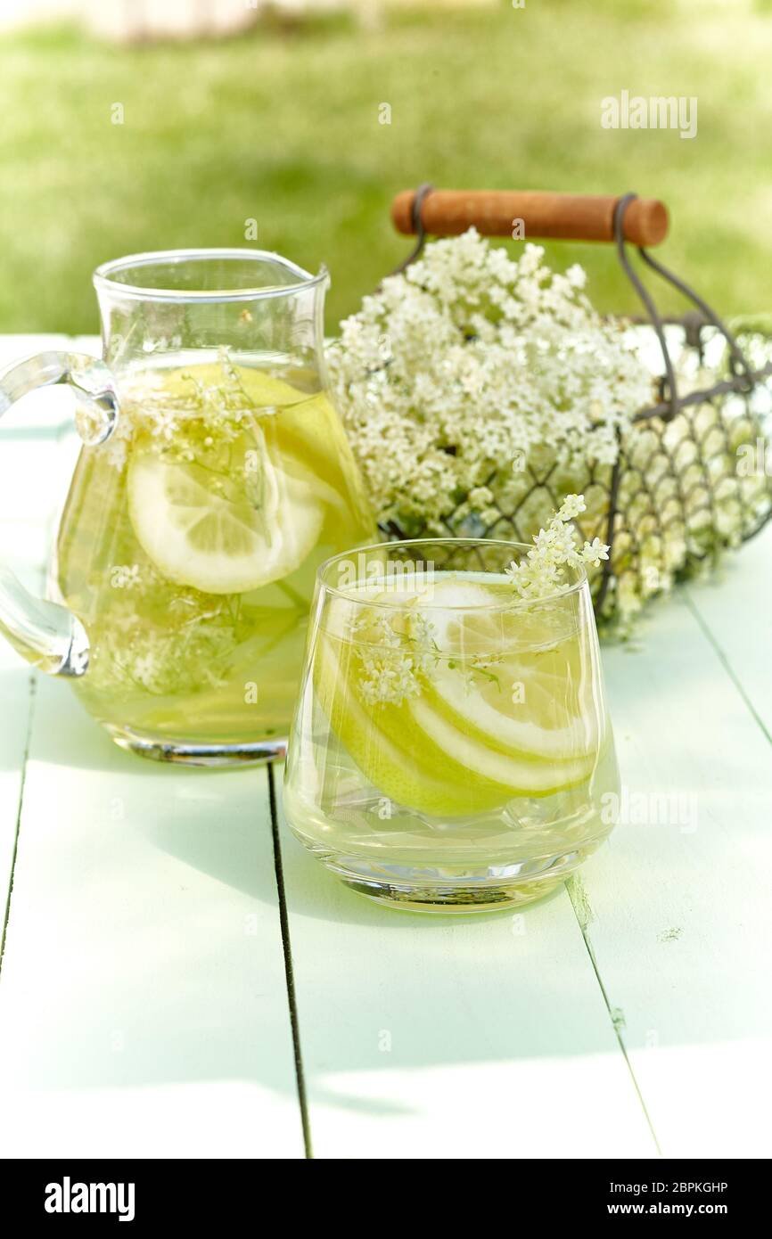 Infusion von frischen elderflowers mit Zitrone in eine Glaskanne und Tumbler draußen auf einer grünen Holz Garten Tisch in einem Konzept von pflanzlichen Heilung serviert, Arom Stockfoto