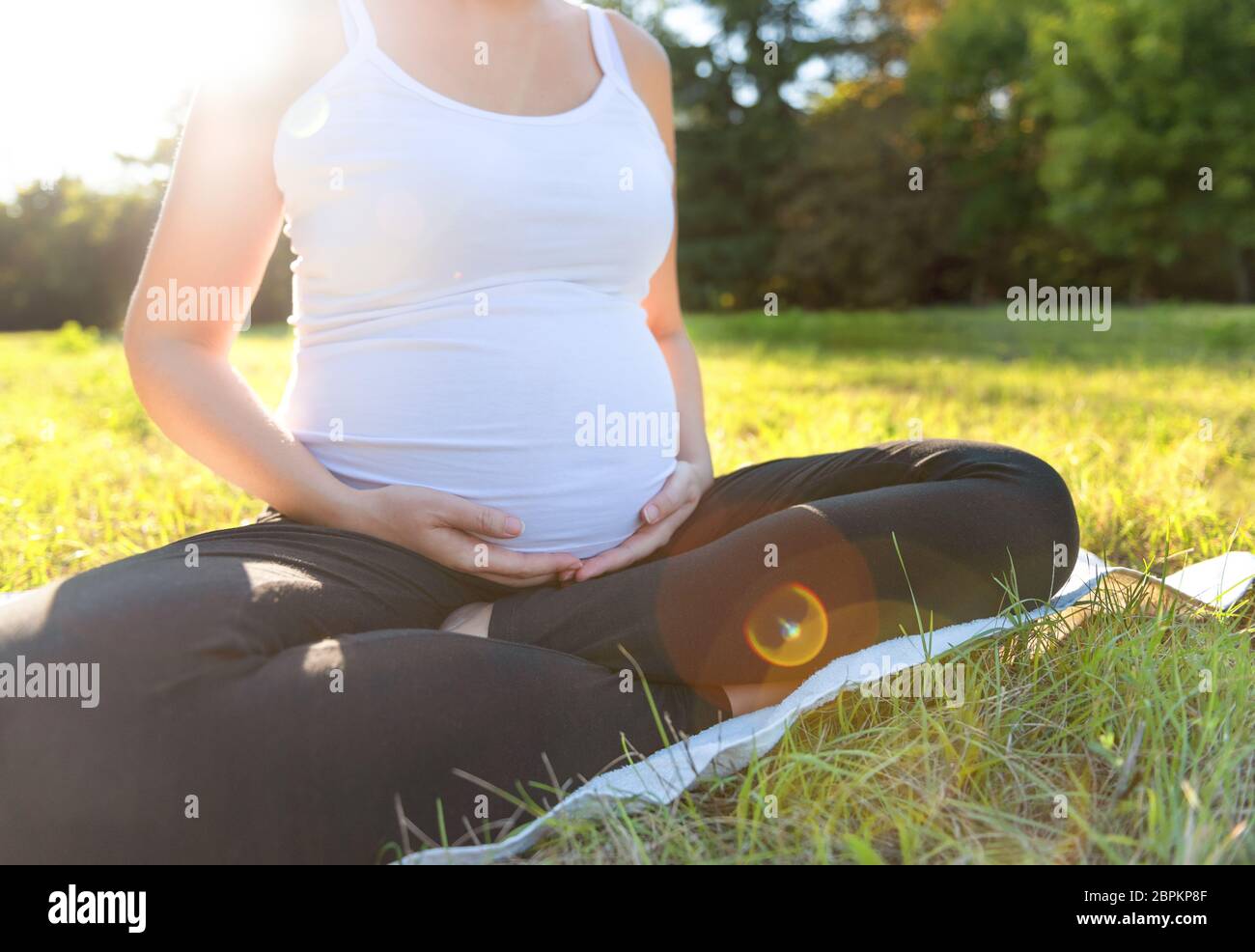 Nahaufnahme einer jungen Frau in Sportkleidung, die während des Trainings im Freien auf einer Übungsmatte sitzt und ihren schwangeren Bauch berührt. Frauen werden erwartet Stockfoto