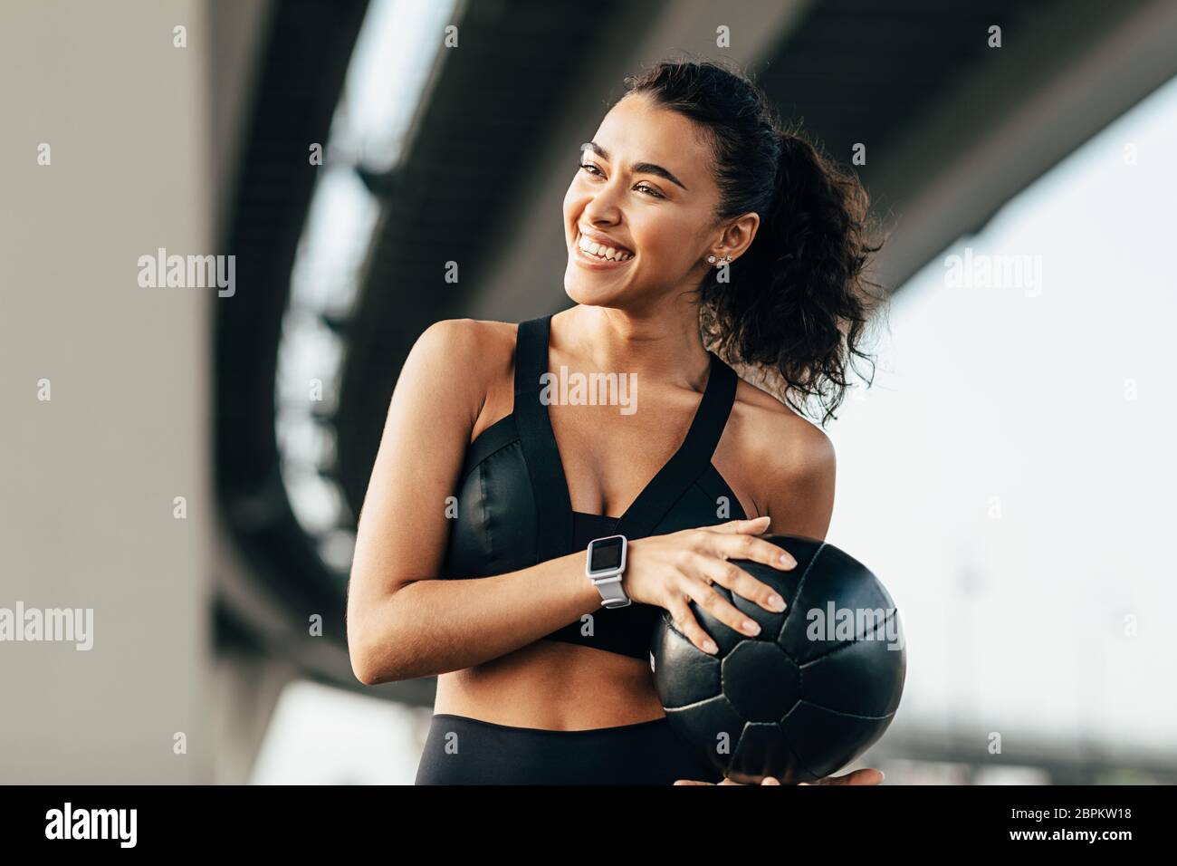 Schöne Frau, die einen Medizinball hält und wegschaut. Lächelnde Sportlerin, die unter einer Autobahn trainiert. Stockfoto