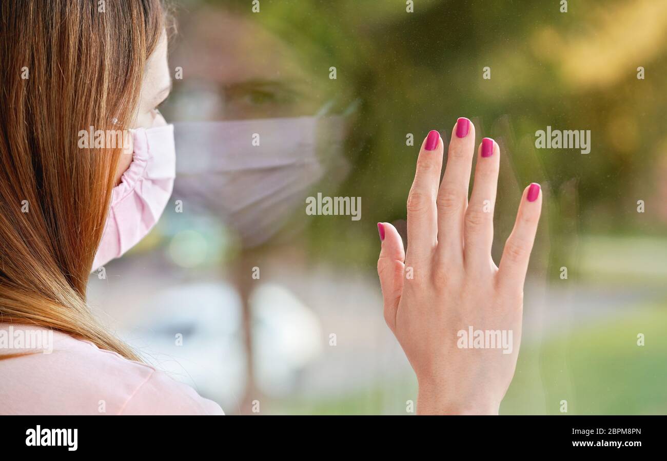 Junge Frau in Virusmaske sieht traurig aus dem Fenster hinter Glasscheibe, berühren sie mit der Hand. Quarantäne oder zu Hause bleiben, um sicher zu sein während der coronavir Stockfoto