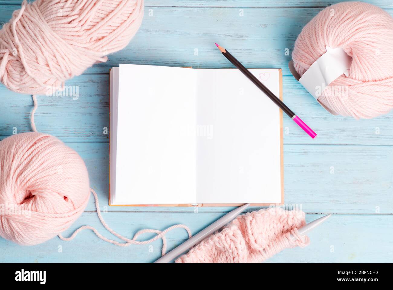 Kreative Kulisse mit pastellrosa farbigen gewundenen Wollen und offenem Notizbuch aus leerem Papier für Notizen und Strickmuster auf hellblauem Holzrücken Stockfoto