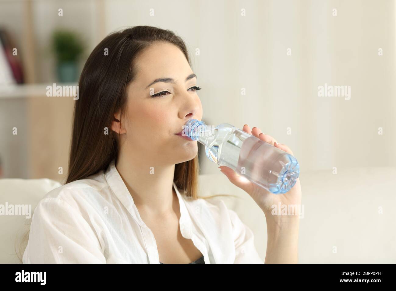 Porträt einer Frau, die Trinkwasser aus der Flasche auf einer Couch im Wohnzimmer zu Hause sitzen Stockfoto