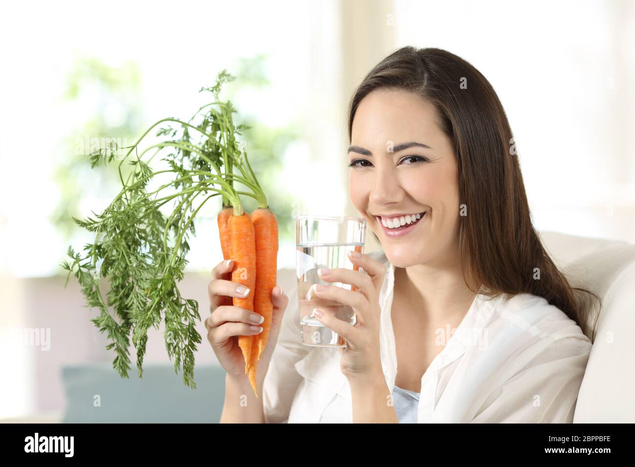 Gerne Frau mit Karotten und ein Wasserglas an der Kamera sitzt auf einer Couch im Wohnzimmer zu Hause suchen Stockfoto