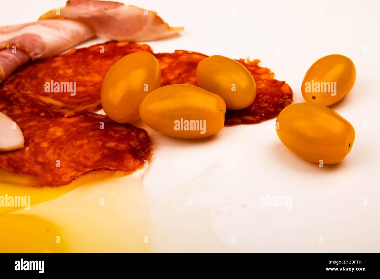 Ein zerbrochenes Hühnereier ohne Schale, Scheiben Wurst und Speck und Tomaten auf weißem Hintergrund. Nahaufnahme Stockfoto