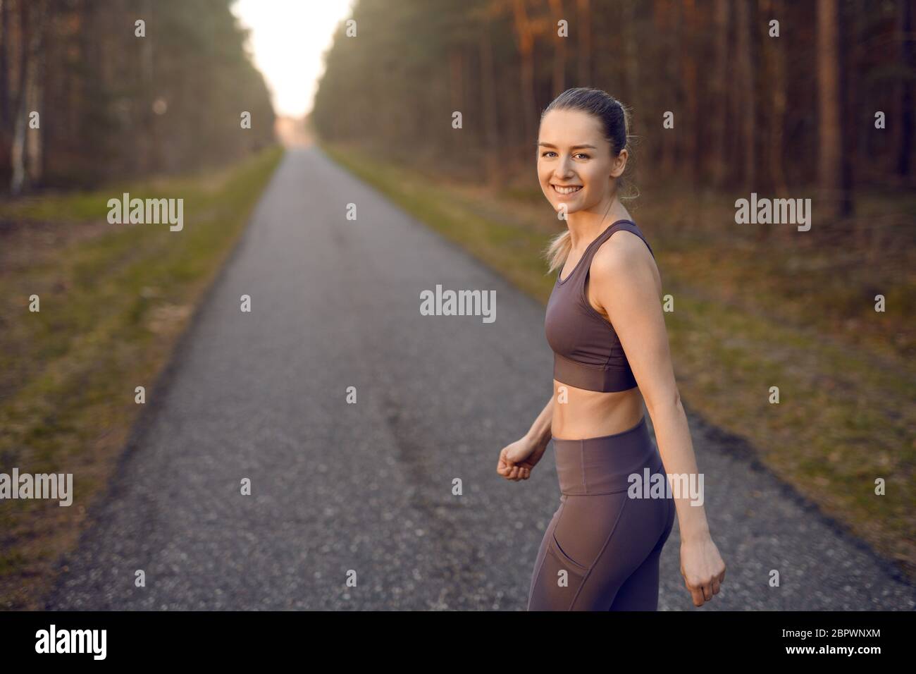 Die junge, athletische Frau passt beim Sonnenaufgang auf einer gerade sich zurückziehenden Straße durch Waldbäume und leuchtet am Ende der Sonne und lächelt Stockfoto