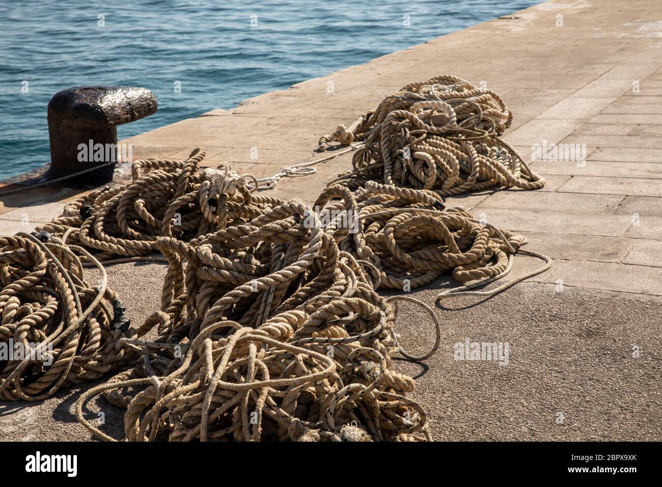 Verankerungsseile auf einem alten Metallpoller und das klare blaue Meer im Hintergrund. Am Hafen liegen viele unterschiedlich große Seile auf dem Boden Stockfoto