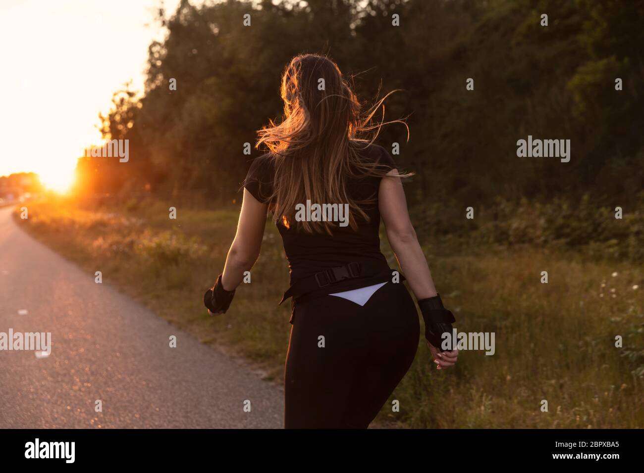 Eine junge Frau Roller Blading auf einer Straße mit Grün im Hintergrund. Sportlich sein, gesund leben. Aufnahme bei Sonnenuntergang, goldene Stunde Stockfoto