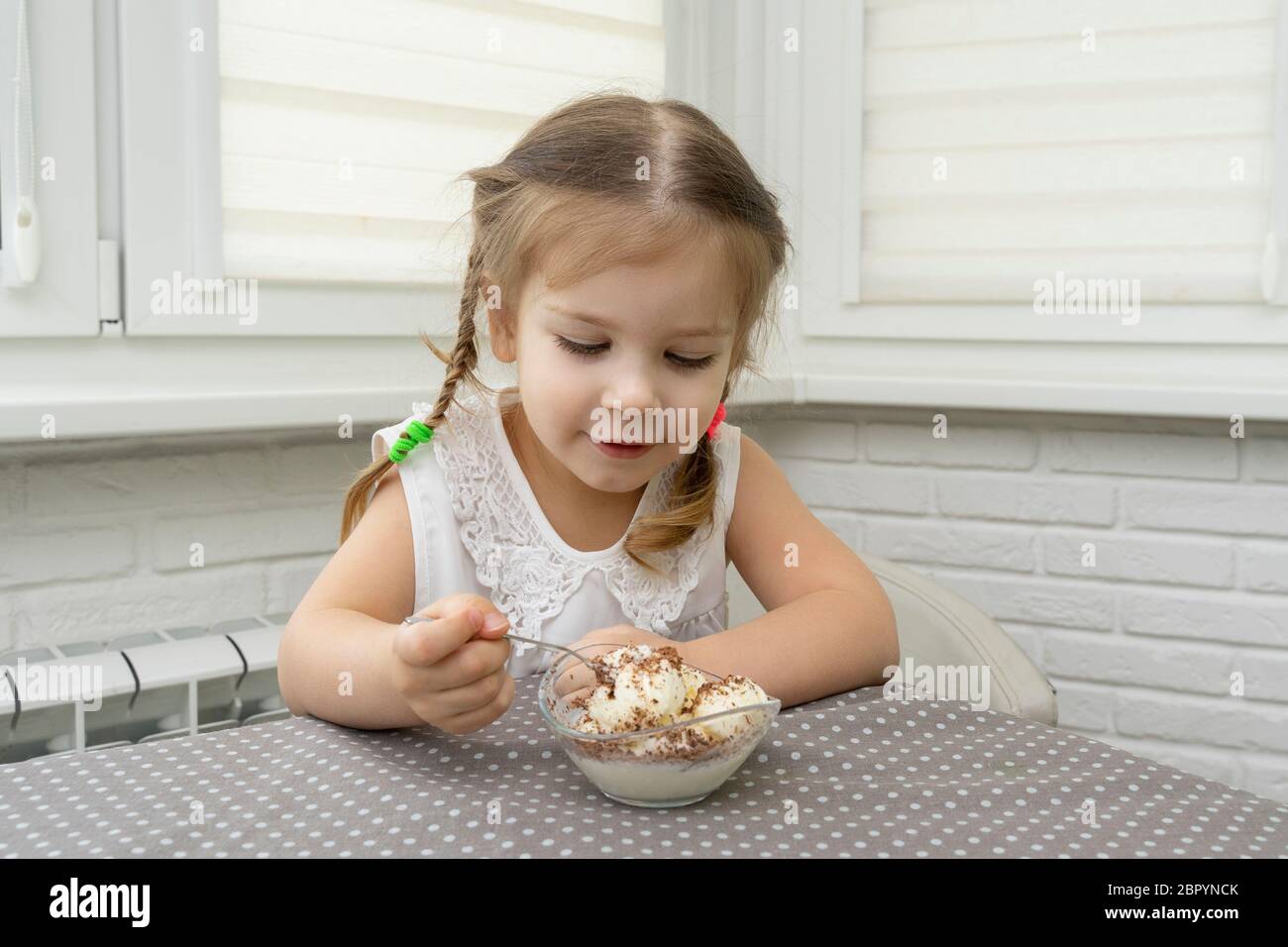 Ein schönes 4-jähriges Mädchen in einer weißen Bluse isst Eis mit Schokolade aus einer Schlangenschüssel an einem Tisch in der Küche mit Vergnügen. Baby ist glücklich ein Stockfoto