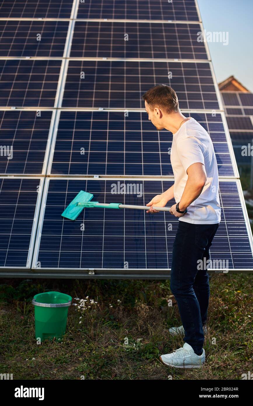 Man reinigt Solarpanel in Solarkraftwerk. Der junge Mann ist stilvoll gekleidet mit einer Uhr an seinen Händen auf einem Grundstück in der Nähe des Hauses Stockfoto