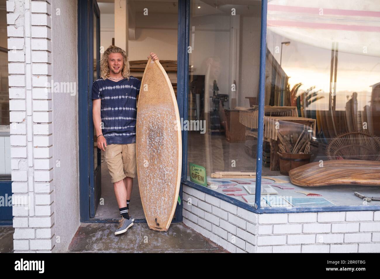 Kaukasischer männlicher Surfbretthersteller, der sich an einer Tür des Eingangs lehnt und ein neues Surfbrett hält und lächelt Stockfoto