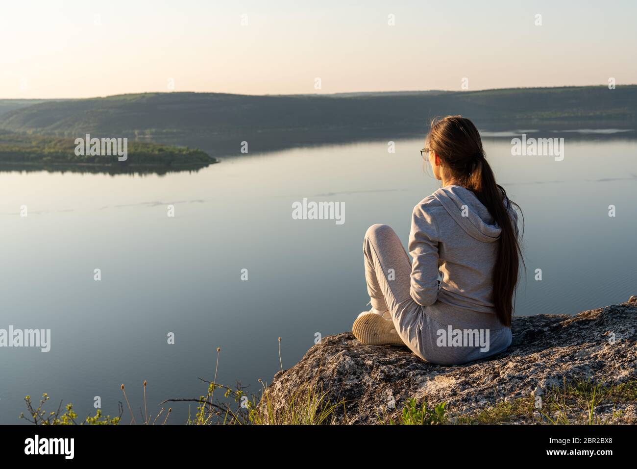 Mädchen sitzt auf einem Felsen, auf einem Berg, schaut in das untere Tal mit einem Fluss Stockfoto