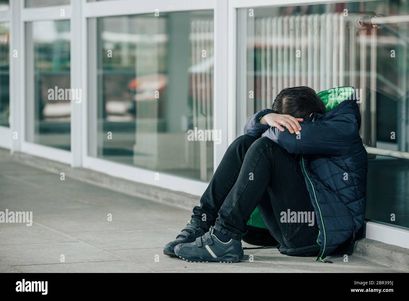 Kleiner Junge oder Kind, der allein auf dem Boden vor der Schule sitzt, nachdem er eine Tat des Mobbgens erlitten hat Stockfoto