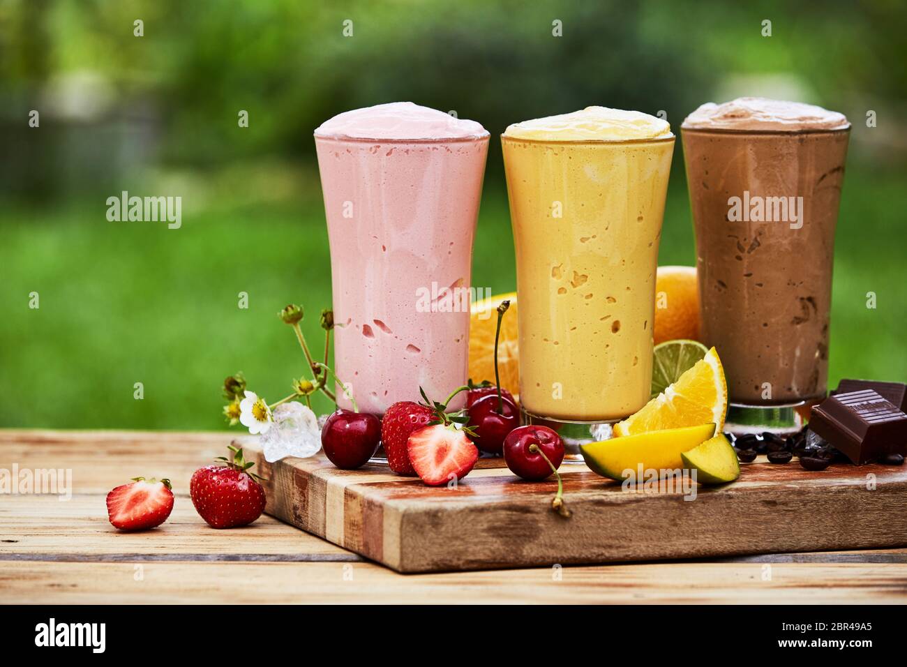 Drei köstlichen frischen tropischen Früchten und Schokolade Milchshakes und Smoothies in hohe Gläser im Freien auf einem Holzbrett serviert im Garten Stockfoto