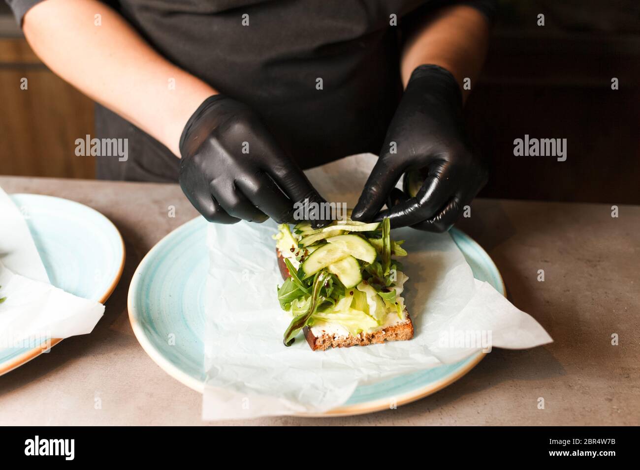 Kochen Sie die Hände Vorbereitung und Herstellung Sandwich. Zubereitung von gesunden vegetarischen Bruschettas in dunklen Handschuhen. Sandwich mit Weichkäse, Rucola und Gurke Stockfoto