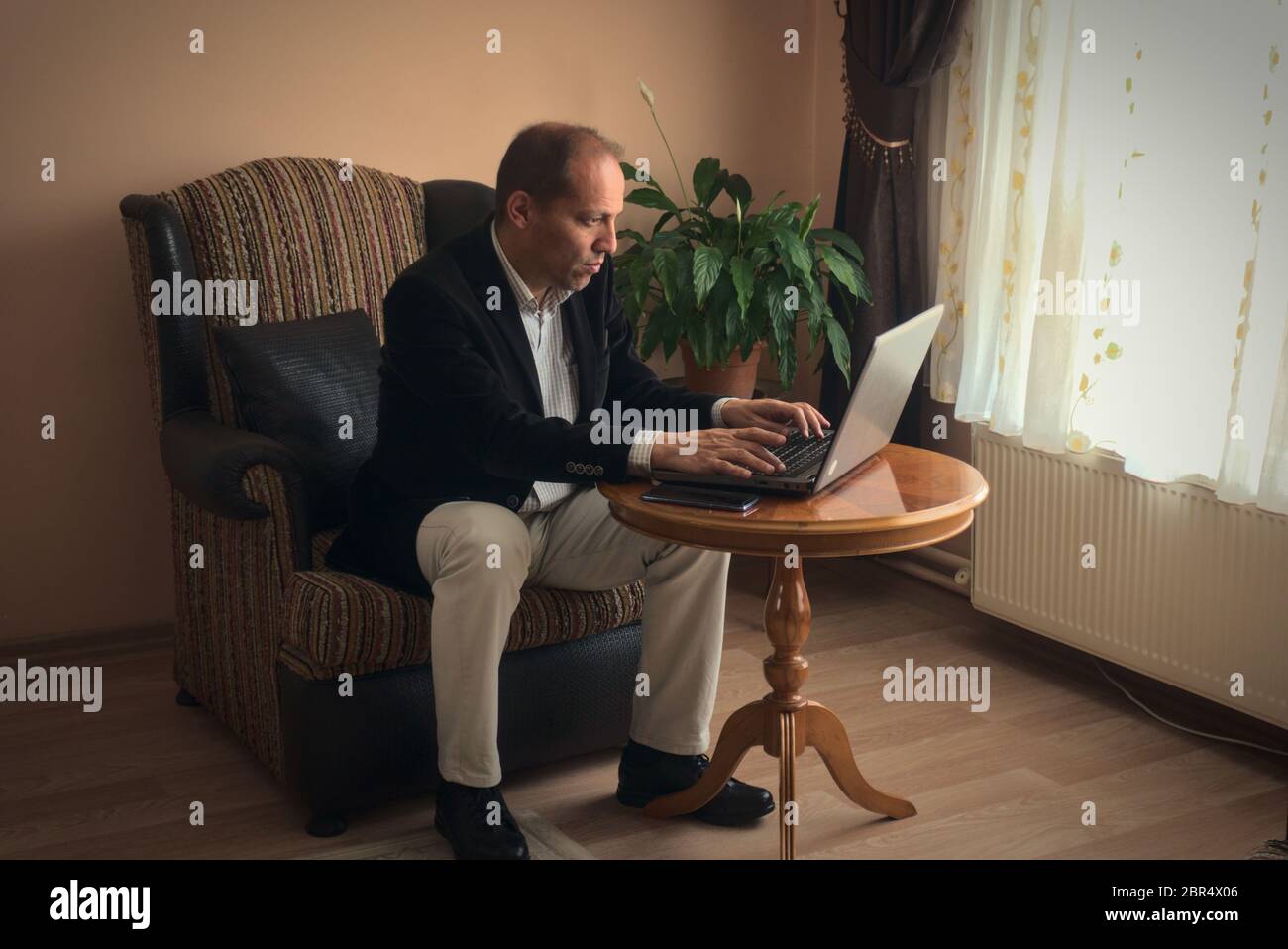 Professioneller und intelligenter Mann mittleren Alters in smarter Kleidung Telearbeit von zu Hause aus, während er auf seinem Sessel sitzt Typen mit seinem Laptop mit einem Pho Stockfoto