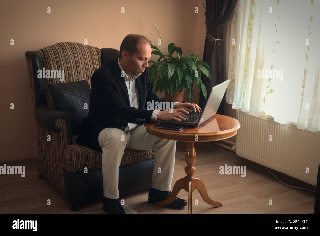 Professioneller und intelligenter Mann mittleren Alters in smarter Kleidung Telearbeit von zu Hause aus, während er auf seinem Sessel sitzt Typen mit seinem Laptop mit einem Pho Stockfoto