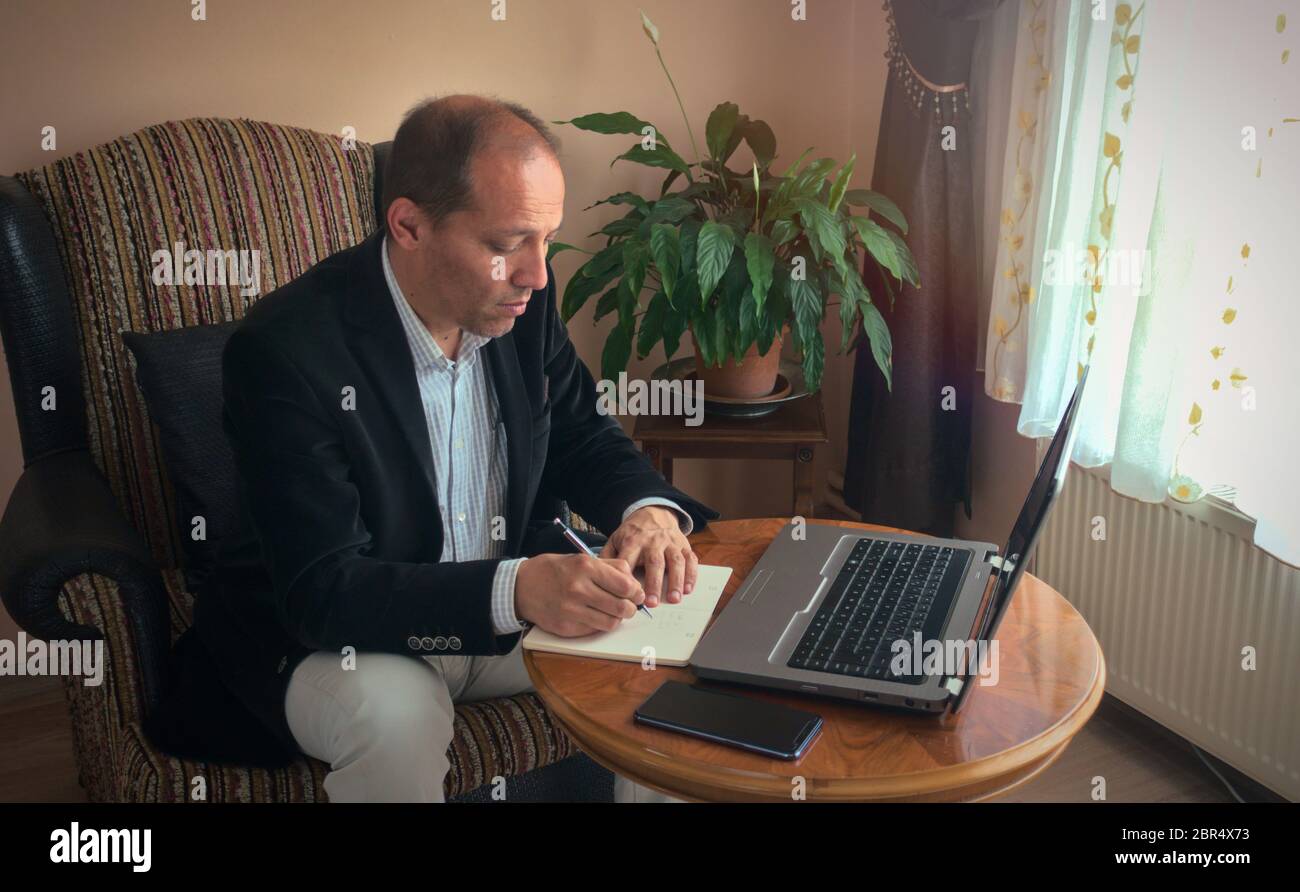 Intelligenter, professioneller Senior, der auf einem Sessel sitzt und von zu Hause aus mit einem Laptop auf einem Tisch arbeitet, der sich in einer Telefonkonferenz Notizen macht, mit einer Anlage auf dem Bac Stockfoto