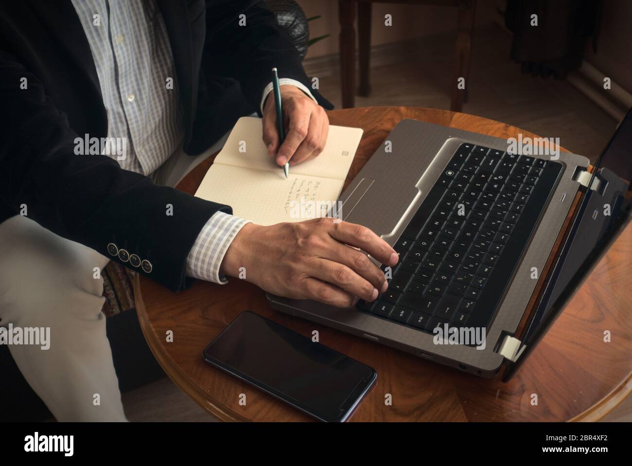 Professioneller Senior, der mit einem Stift Notizen macht, während er mit seinem Laptop eine Konferenz führt und ein Telefon daneben auf einem Tisch steht Stockfoto