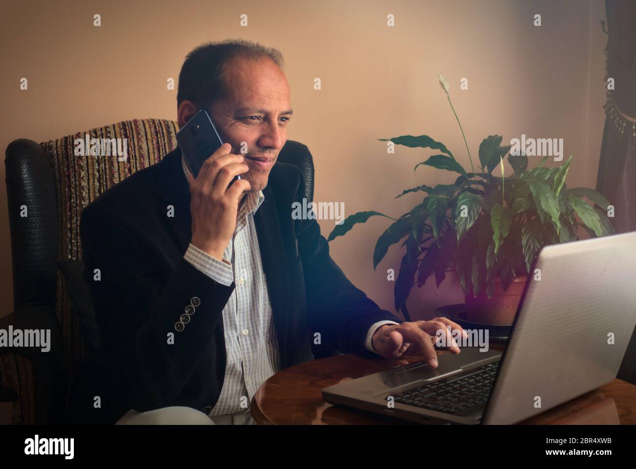 Glücklich und lächelnd, sitzt auf dem Sessel und telefonert mit einem Freund, während er mit seinem Laptop im Internet surft, zu Hause, Seitenansicht, Blick Stockfoto