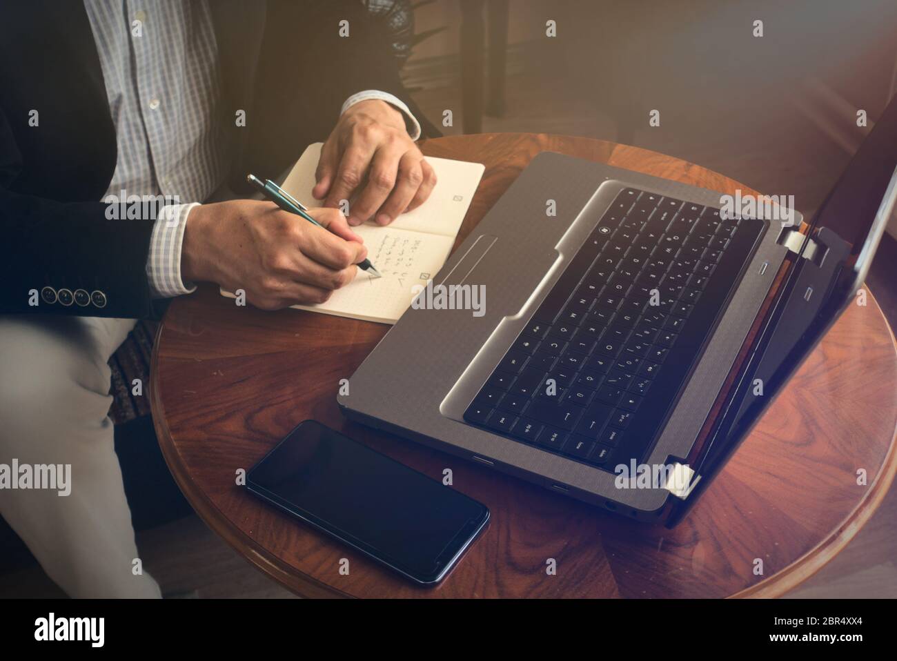 Professioneller Senior, der mit einem Stift Notizen macht, während er mit seinem Laptop eine Konferenz führt und dabei ein Telefon auf einem Tisch daneben hat Stockfoto