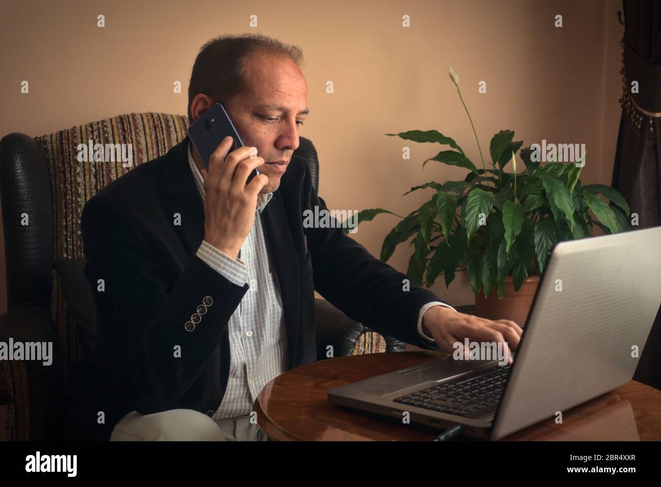 Intelligent, professionell, ängstlich Senior Mann in smarter Kleidung sitzt auf seinem Sessel telefonieren mit jemandem, während die Arbeit mit seinem Laptop, mit einem Plan Stockfoto