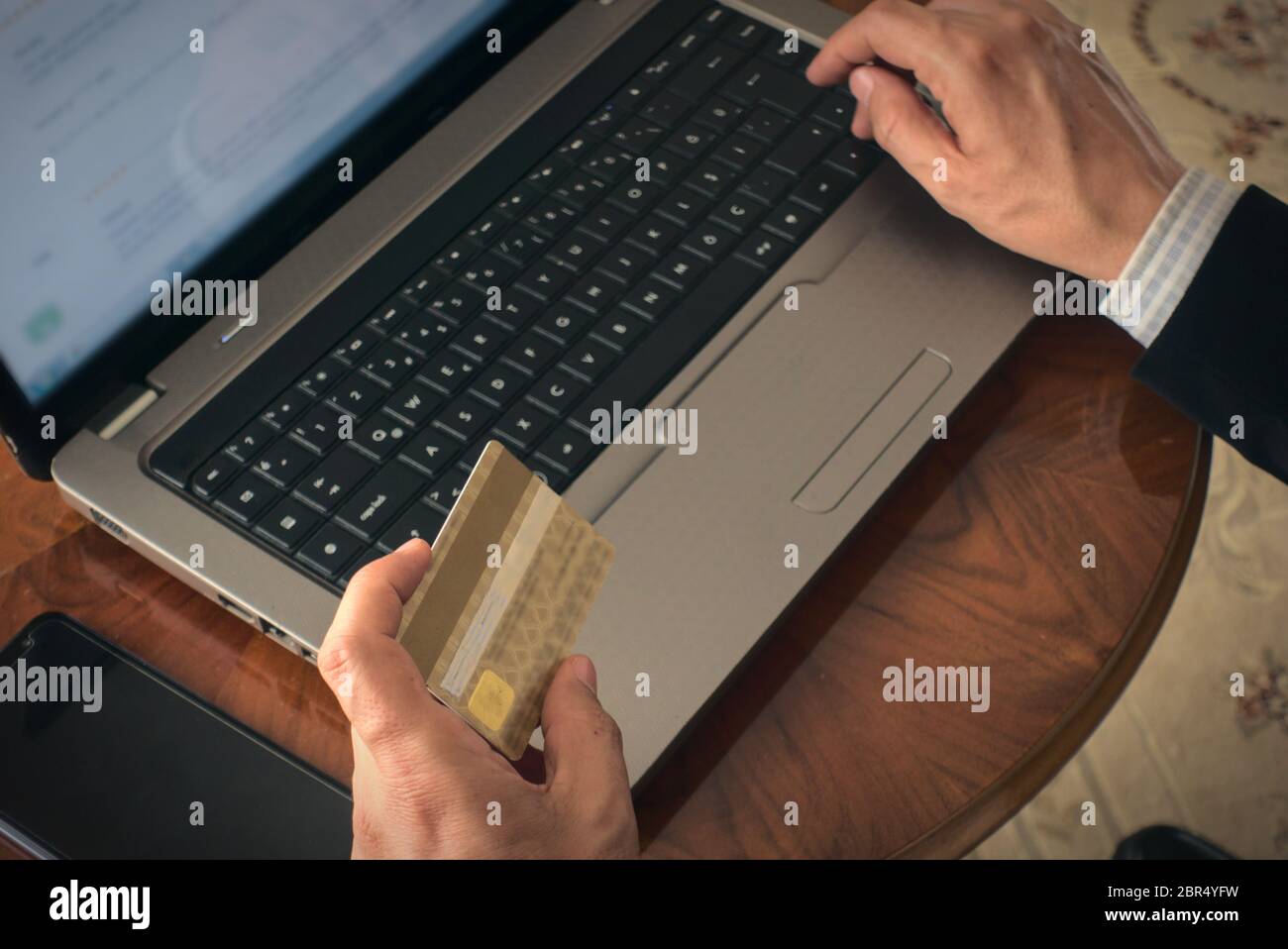 Nahaufnahme Mann hält eine Kreditkarte Online-Shopping von seinem Laptop mit einem Telefon daneben auf einem Tisch zu Hause Stockfoto